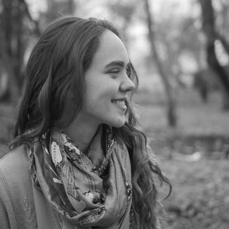 A woman smiles while looking to the right. She has dark, curly hair and is wearing a scarf. In the background, it is winter.