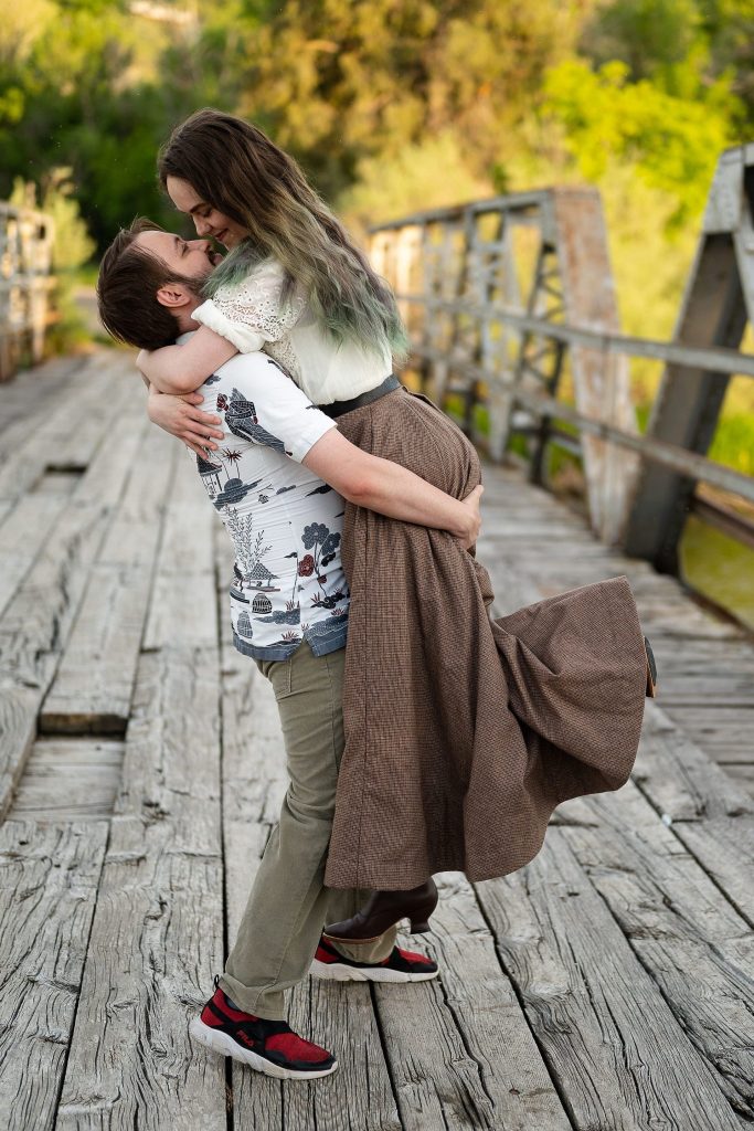 A man holding a woman on a bridge. Both are smiling. The woman is wearing a floor-length victorian-style skirt made from tweed. 
