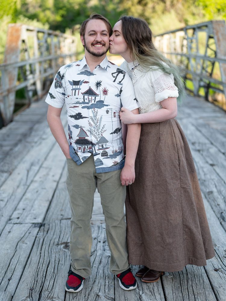 A man holding a woman on a bridge. Both are smiling. The woman is wearing a floor-length victorian-style skirt made from tweed. 