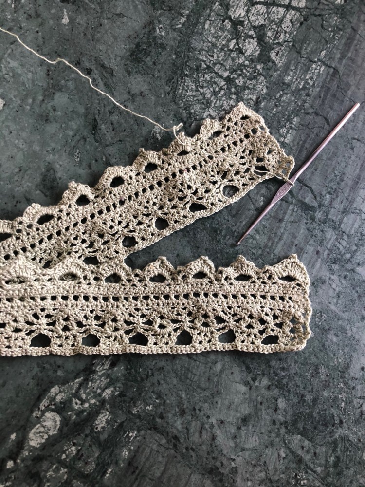 Lace trim lying on a marble table. One end of the lace is raw, still attached to a small metal crochet hook.
